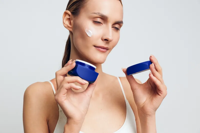 Young woman applies face cream on cheek holds open jar of moisturizer has clean fresh skin closes eyes enjoys skincare routine isolated on light gray background.