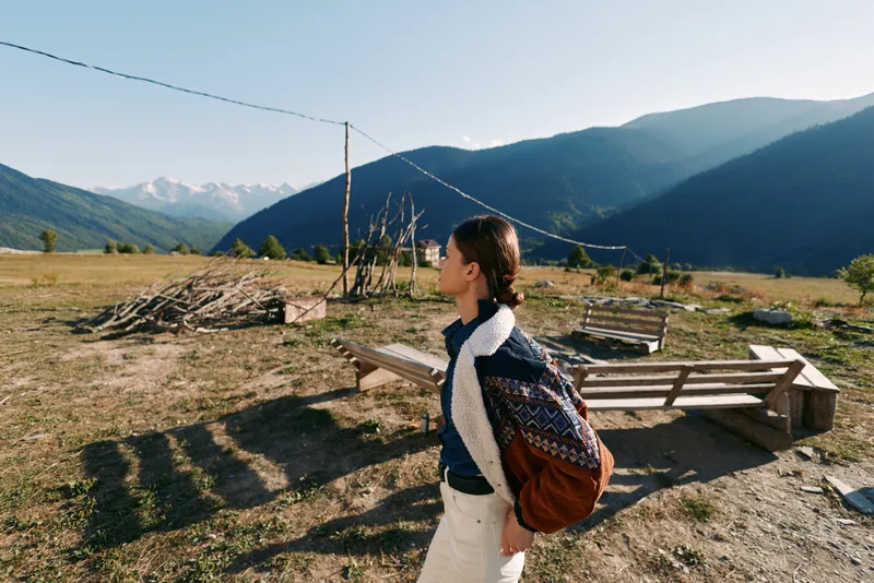 Woman with backpack hiking across a rural meadow near mountains, outdoor travel portrait of young traveler in sweater walking past wooden benches and fences under clear blue sky.