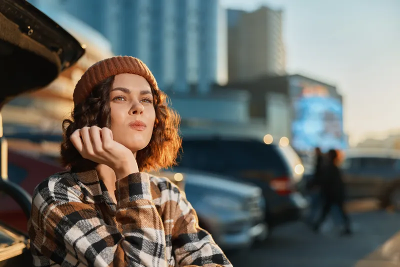 Woman portrait in urban street with thoughtful gaze, candid authenticity and golden glow at sunset, beanie and plaid jacket evoke mindful living and emotional storytelling.