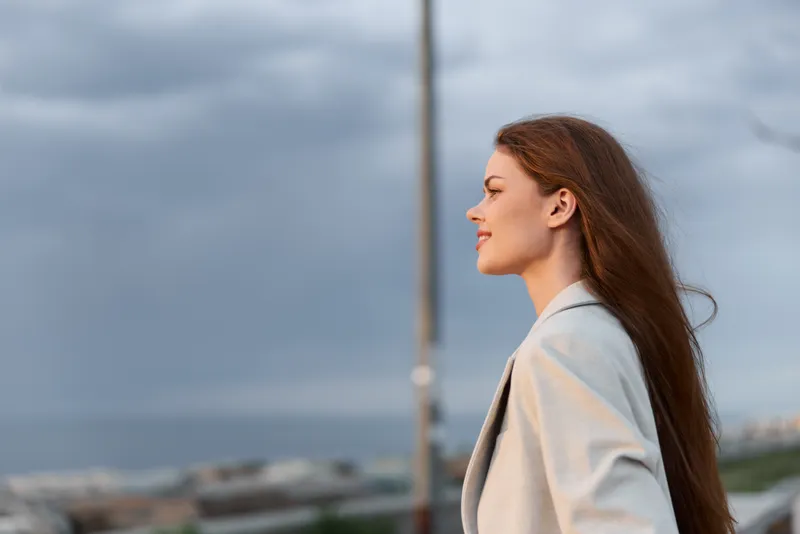 Confident business woman with long hair in a stylish blazer against a cloudy sky, embodying professionalism and ambition in a dynamic outdoor setting.