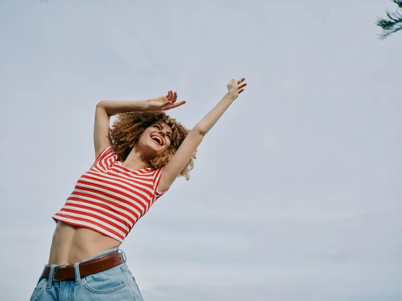 Joyful young woman with curly hair wearing a red striped crop top and jeans, arms raised against a bright sky, capturing carefree summer energy and movement.