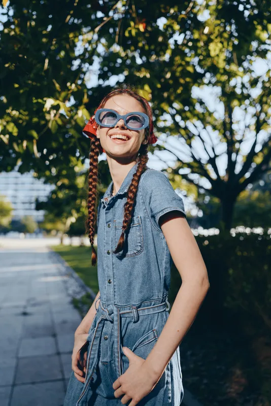 lifestyle woman in modern boho-western denim street style smiling joyfully outdoors with film color tone, sunglasses, and braided hair under bright sunlight