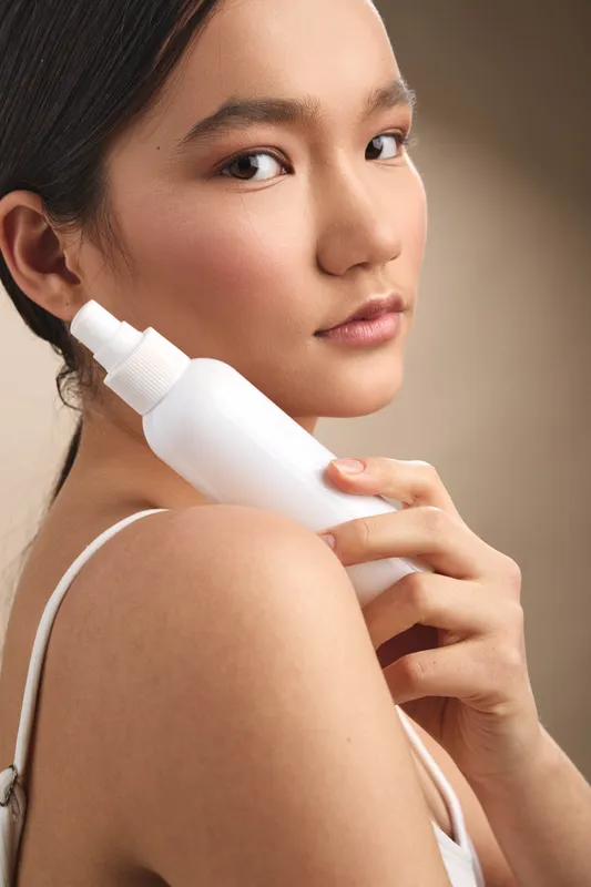 Asian woman holds a white skincare spray bottle against her shoulder, showcasing clear skin and soft makeup, exuding calm confidence and a focused beauty routine.