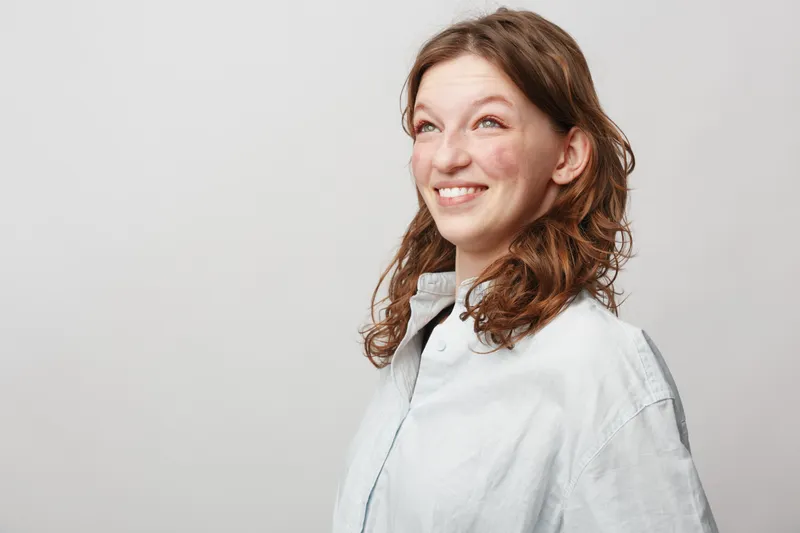 Smiling young woman with curly brown hair looks up with bright eyes wearing a light shirt. Studio portrait on white background with natural expression and casual style.
