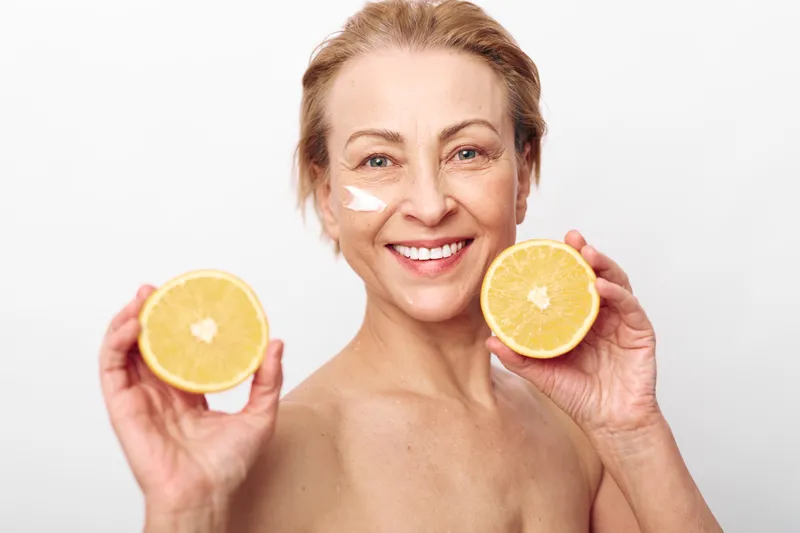 Smiling mature woman with short hair holding two halves of a fresh grapefruit near face, showing healthy skin and natural beauty. Concept of skincare, vitamin c, and wellness in studio.