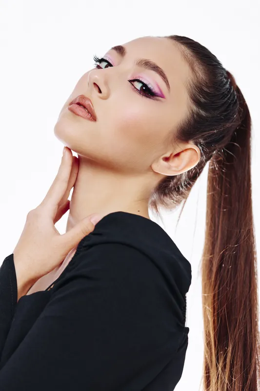 Young woman with creative pink eyeshadow and sophisticated neutral lipstick poses with her hand near neck. Fashion beauty theme on white background focuses on makeup and style.