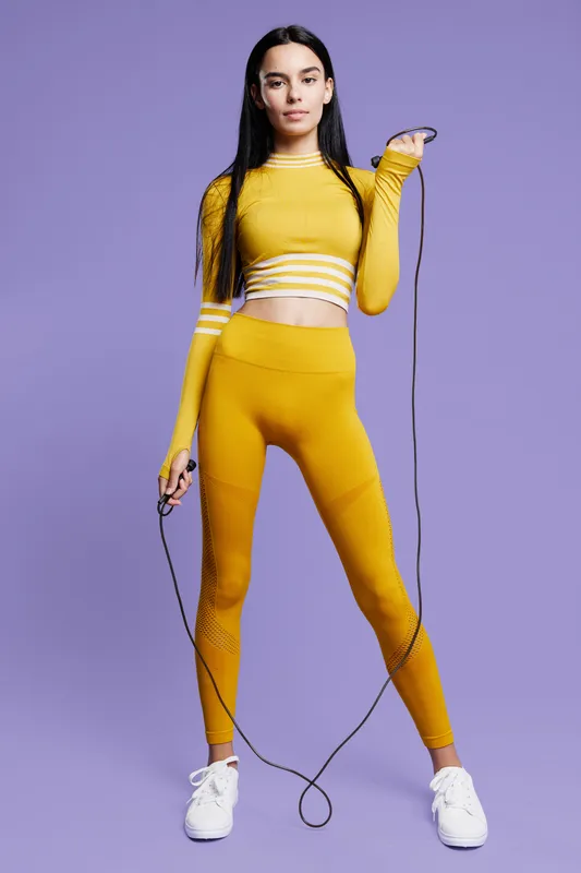 Woman sport fitness wearing yellow activewear holding jump rope on purple background, showcasing health, exercise, and strength in a modern fitness setting.
