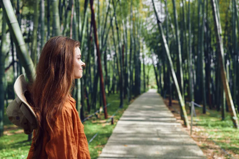 woman profile redhead hat bamboo path in a green forest walkway. Young woman in orange jacket stands on trail, enjoying sunlight and serene nature scene during relaxed outdoor travel.