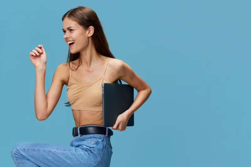 Smiling woman holding laptop, dressed in casual beige top and blue jeans, posing joyfully against a plain blue background with vibrant energy.