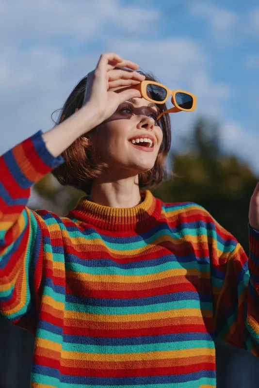 Woman striped sweater yellow sunglasses smiling short hair outdoors looking up in a sunny park, casual outfit and cozy knitwear for lifestyle, travel and joyful fashion portrait.