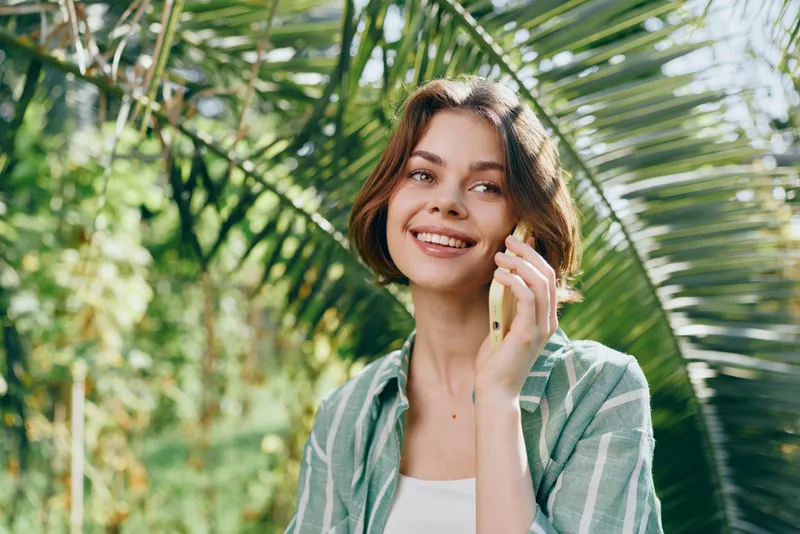 Young woman in casual attire speaks on a mobile phone outdoors, surrounded by lush green foliage and bright sunlight, with a warm smile.