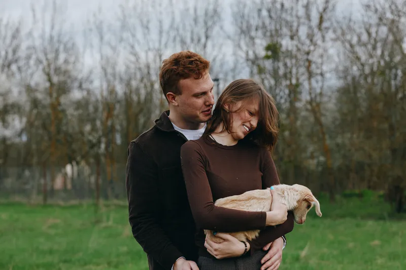 A young couple outdoors in a park cradle a small goat kid, sharing a warm moment of affection. The scene captures rural lifestyle, bonding, and gentle animal companionship.
