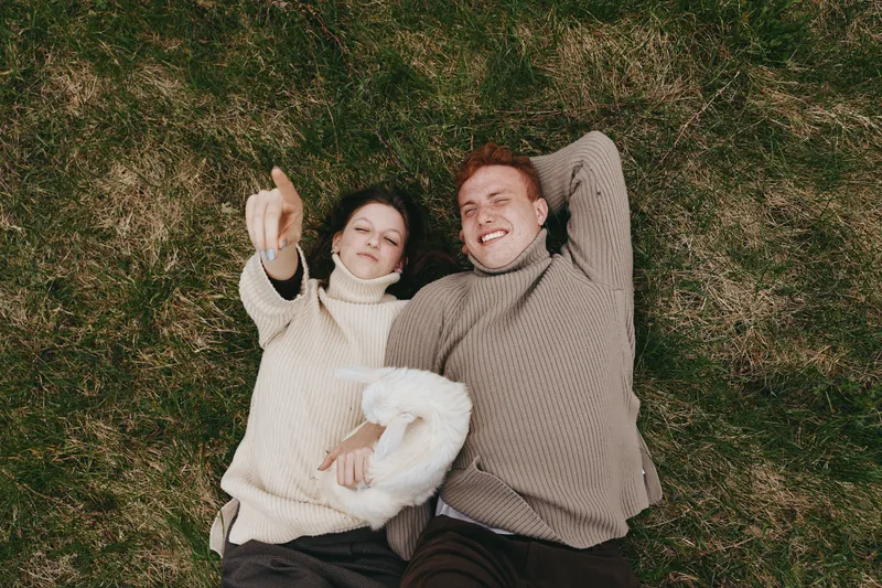 Couple outdoors, lifestyle moment, grass backdrop, sweater wearers, cozy, smiling, relaxed, daylight ambiance, candid connection, warm natural colors