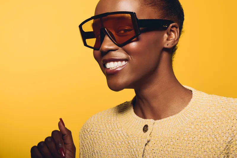 Smiling African American woman with dark skin wearing oversized sunglasses and a yellow sweater poses against a bright yellow background. Concept of fashion, confidence, and style.