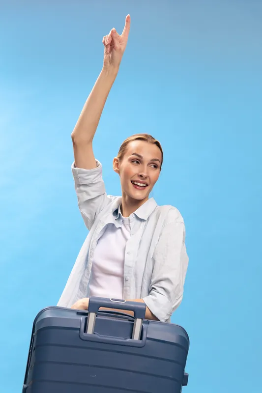 Happy young woman with suitcase raises hand and smiles, ready for travel. Casual outfit and blue background create cheerful concept of vacation, trip, and adventure for tourism.
