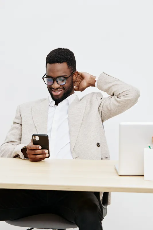 Confident young African-American man wearing glasses and a beige blazer, smiling while looking at his smartphone during work or leisure at a modern office desk with a white background. People lifestyl