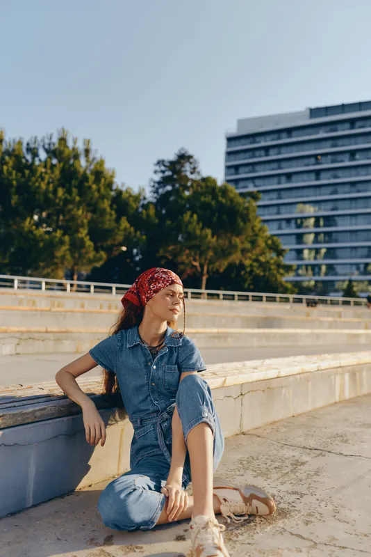 young woman sitting outdoors in casual denim outfit with red headscarf on sunny day, relaxed mood and urban background