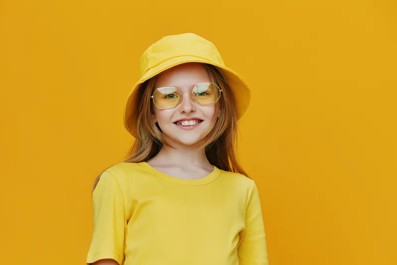 Cheerful girl in yellow outfit and bucket hat against a vibrant yellow background, exuding happiness and summer vibes, perfect for children s fashion content
