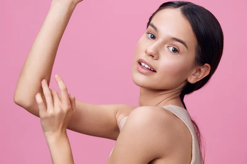 A young woman with smooth skin showcasing her arm in a minimalistic setting, conveying beauty and confidence against a pink background, ideal for wellness and skincare themes