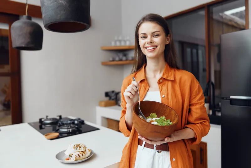 Веселая женщина демонстрирует вкусный салат на стильной кухне, пропагандируя здоровое питание и яркий образ жизни со свежими ингредиентами и теплыми цветами