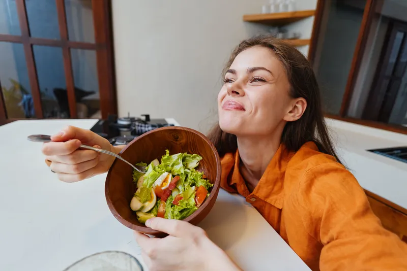 Счастливая молодая женщина наслаждается вкусным салатом на своей стильной кухне, пропагандируя радость здорового питания с помощью ярких цветов и природных элементов