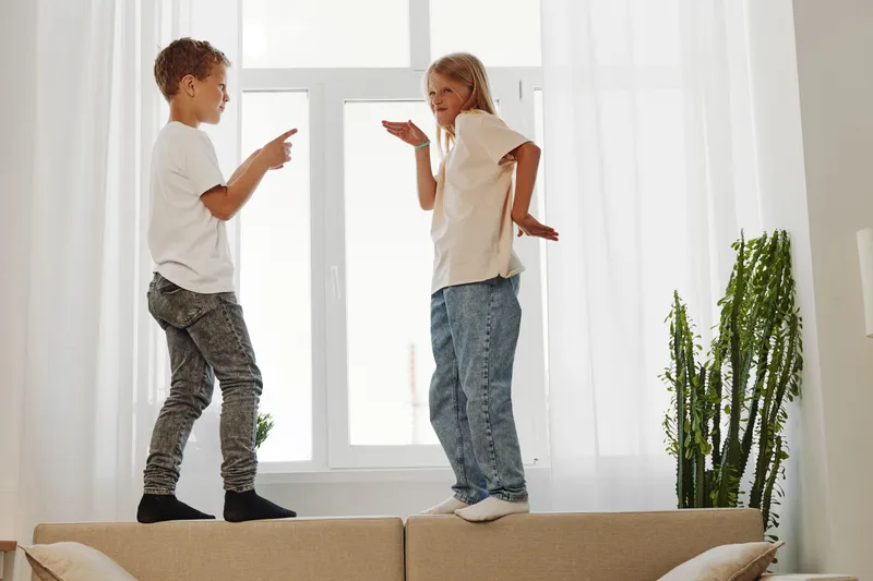 Two children standing on sofa near window playing and laughing in bright room, casual clothes and joyful mood. Concept of childhood fun, home activities, sibling interaction.