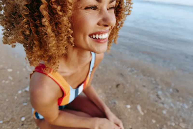 Curly haired woman smiling on the beach, wearing a colorful swimsuit, warm sunlight lighting her face, relaxed vacation mood, close up portrait near shoreline, joyful summer freedom