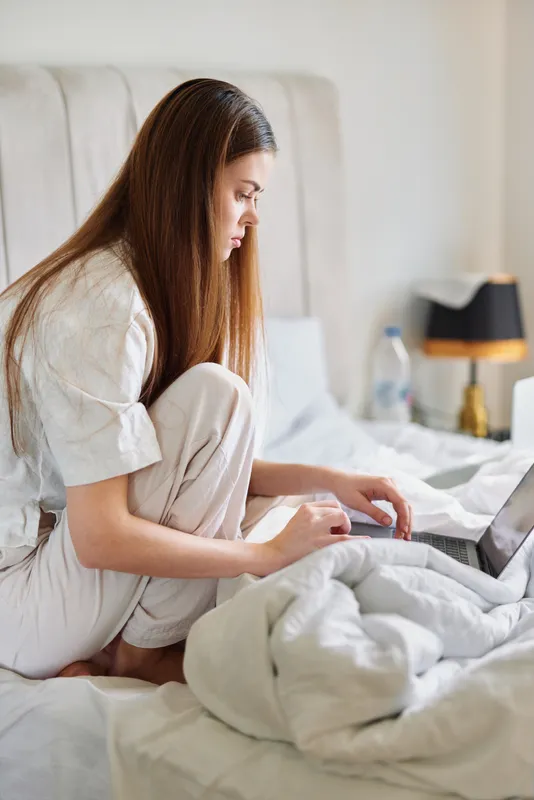Woman on bed in pajamas working on laptop in bright bedroom, morning routine and remote work scene with cozy sheets, focused young adult using computer for study or job at home.