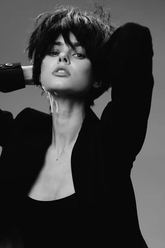 Black and white portrait of a stylish woman with short hair in a blazer posing confidently with raised arms.