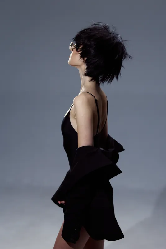 Profile of a stylish woman with short black hair wearing black clothing and sunglasses in a modern minimalistic setting.