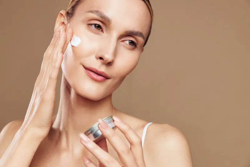 Woman applying moisturizer cream to face, beauty skincare routine, soft spa mood, gentle smile, natural skin texture, bright studio light, smooth beige background, wellness care
