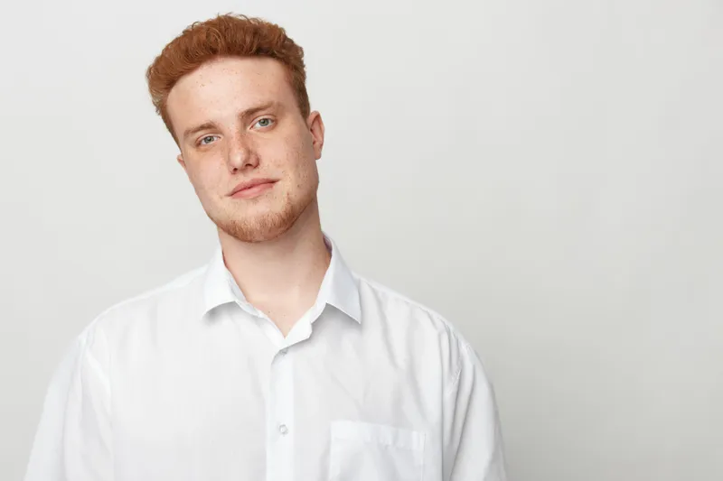 Portrait of a young man with red hair wearing a white shirt against a plain light background. Casual style, confident expression, and natural lighting in studio.