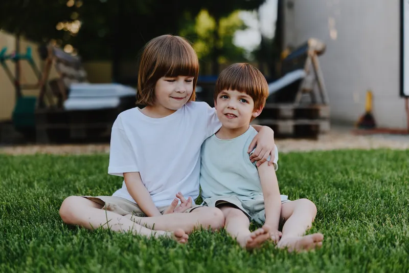 Two cheerful boys sitting together on green grass, enjoying a sunny day, sharing a moment of joy and friendship in a relaxed outdoor setting.