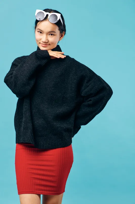 Young woman in black sweater and red skirt posing confidently with sunglasses on head against blue background. Fashionable casual outfit and modern style standing with hand near face.