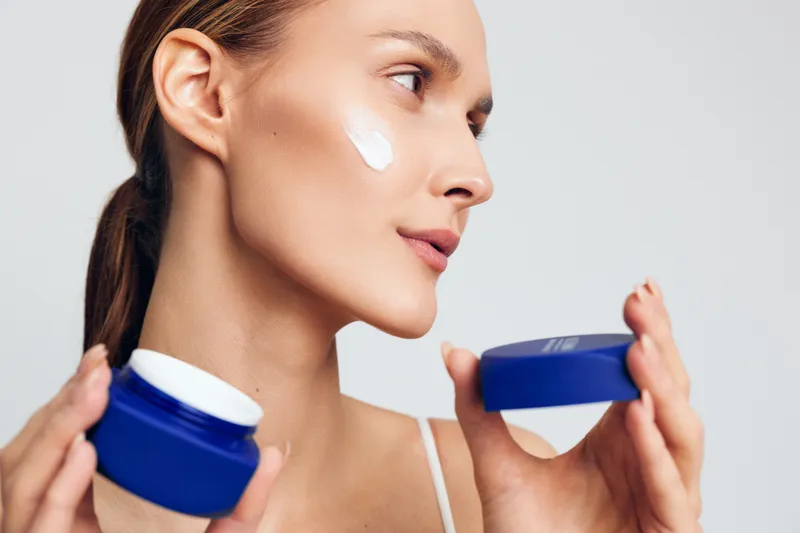 Young woman applying moisturizing cream on her cheek holding a blue jar with white cream on a white background. Concept of skincare, beauty, and daily facial care routine.