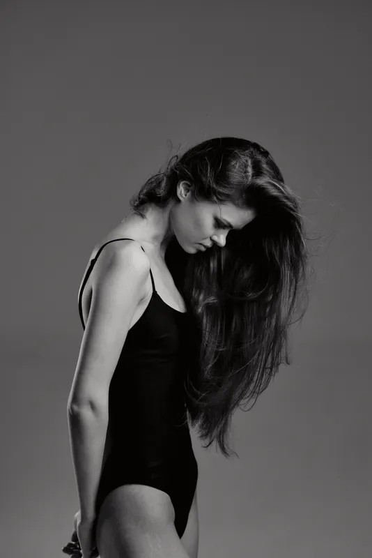 Black and white portrait of a contemplative woman with long hair wearing a black bodysuit standing against gray background