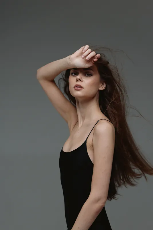 Young woman with long hair wearing black dress posing gracefully with hand on forehead against gray background.