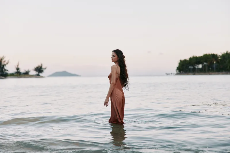 A woman stands waist-deep in a calm sea at dusk, wearing a flowing dress, as gentle waves kiss a tranquil shore and distant trees glow softly.