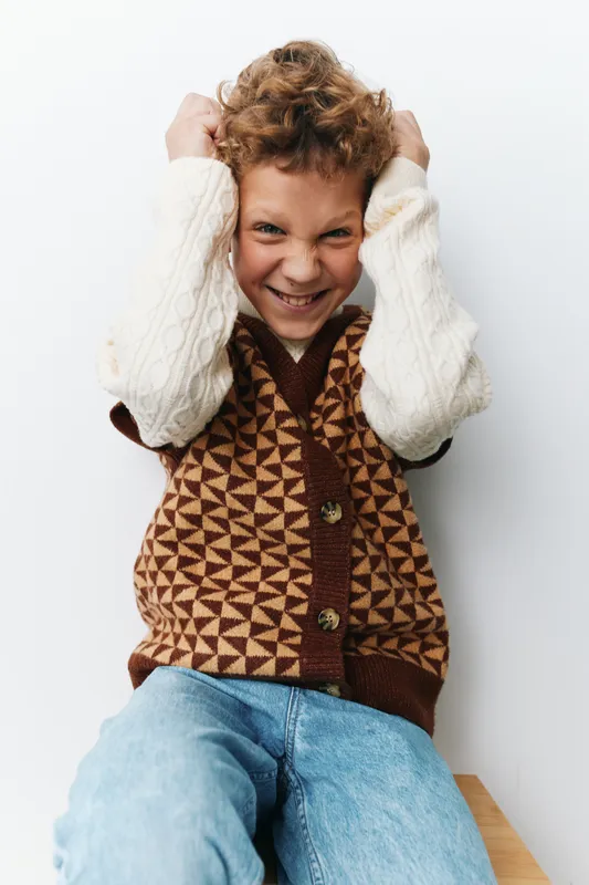 Playful boy grins with hands in hair, wearing cream knit sweater vest and denim jeans, posed against a clean white background with cheerful energy.