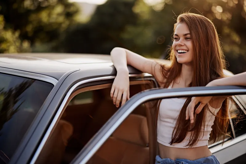 Smiling young woman leaning on a car, enjoying a sunny day, wearing casual summer clothes, and surrounded by a lush green background.