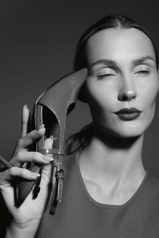 Closeup portrait of a white woman with fair skin holding high heel shoe near face. Concept of fashion, beauty, and style in black and white photography.