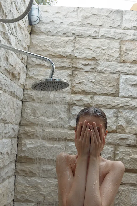 naked woman washing face under outdoor rain shower on stone wall background, natural skin care, refreshing water droplets, morning routine, peaceful atmosphere