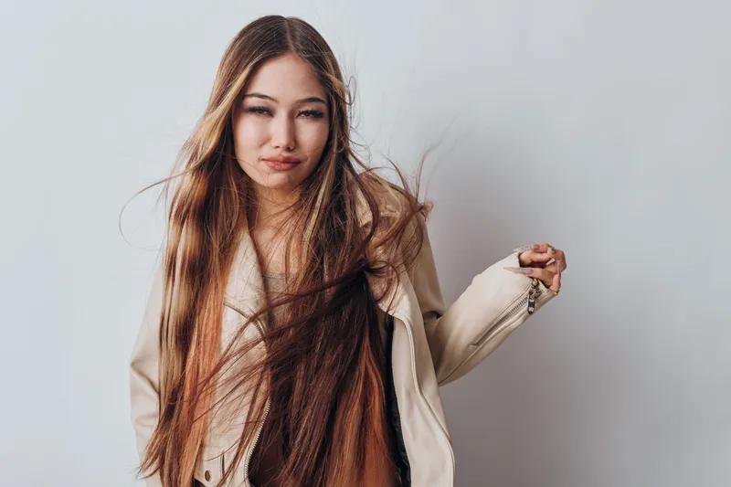 Young woman with long hair wearing beige coat standing against gray background looking at camera with slight smile and casual pose for fashion and beauty concept.