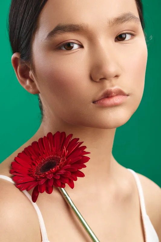 Asian woman with a red flower resting near her shoulder against a vibrant green backdrop, presenting a serene, elegant portrait with soft lighting and calm mood.