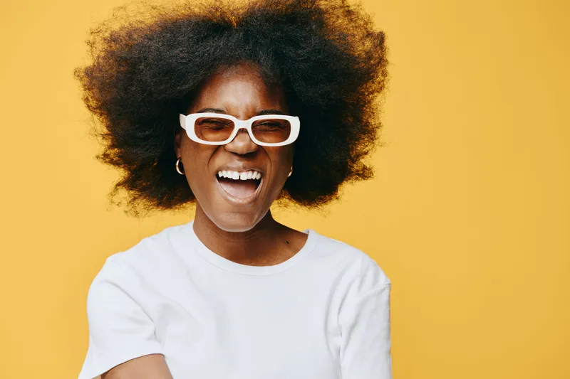 Smiling African woman with curly hair, wearing glasses and a white shirt, vibrant yellow background exuding joy and positivity