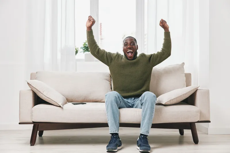 Excited African American man celebrating at home, wearing a green sweater and jeans, on a cozy couch in a bright room with a joyful expression