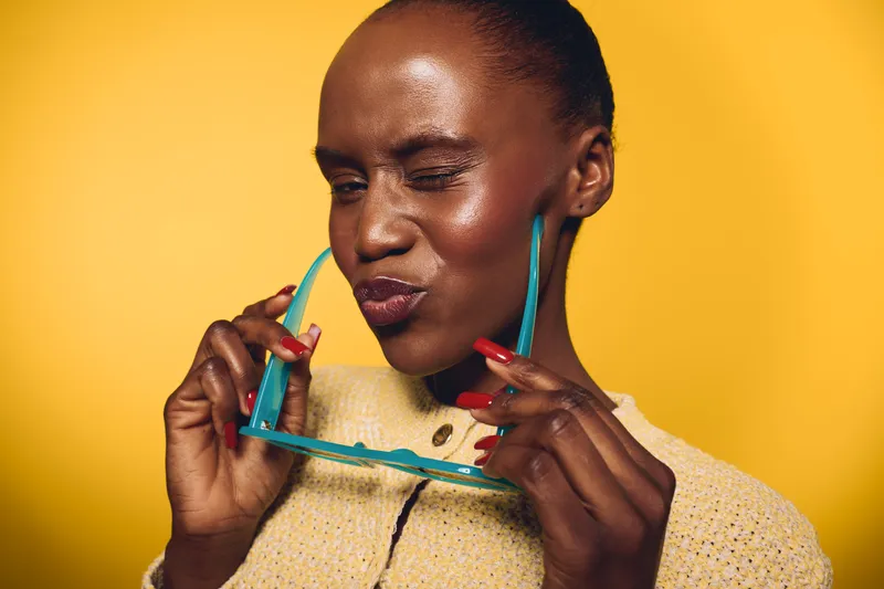 Young African woman with dark skin winks holding blue sunglasses, wearing yellow sweater on yellow background. Concept of fashion, beauty, and playful mood with stylish accessories.
