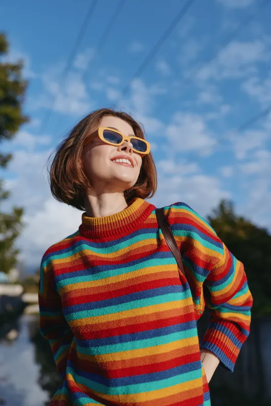 Woman in her late 20s wearing a colorful striped sweater and retro sunglasses, short hair bob, smiling outdoors on a sunny urban street with shoulder bag for lifestyle travel.