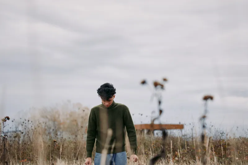A lone young man in a green sweater stands in a windswept field, captured in a genuine documentary style that emphasizes true-to-life emotion, unposed presence and natural detail.