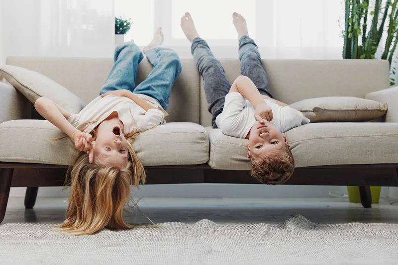 Two happy children lie upside down on a beige sofa in a bright living room, laughing and having fun together. Concept of childhood joy, friendship, and home comfort.